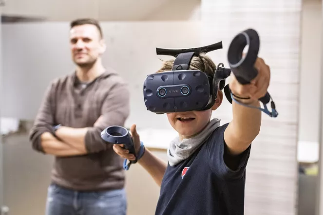 Ein Junge trägt eine VR-Brille und spielt, dahinter steht ein Pädagoge und schaut zu.