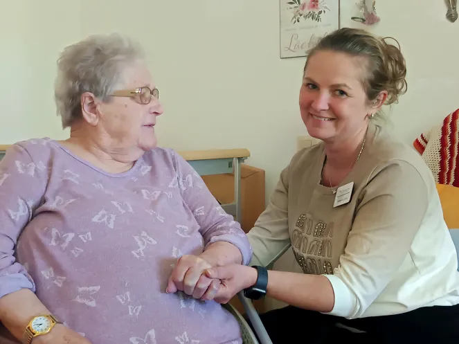 Pflegefachkraft hält die Hand einer Seniorin im Rollstuhl und lächelt