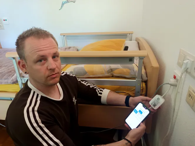 Ein Mann hockt vor einem Krankenbett und hält ein Handy in der Hand.