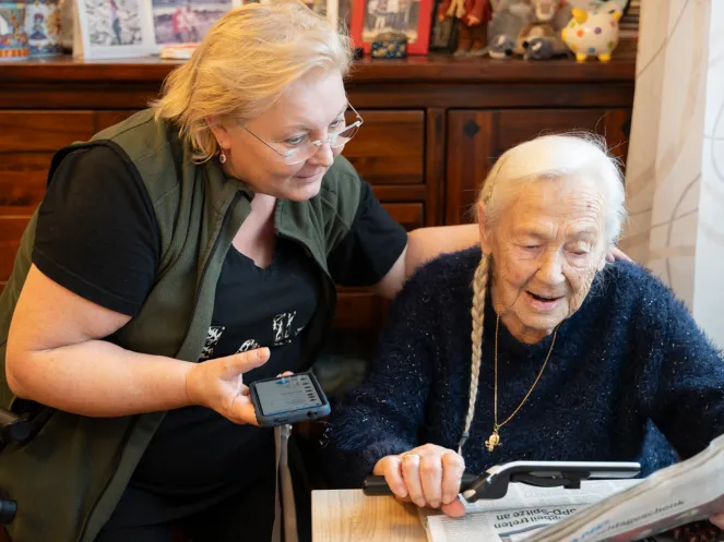 Eine Pflegerin legt den Arm um eine Seniorin, die Zeitung liest. Die Pflegerin hält ein Handy in der Hand.