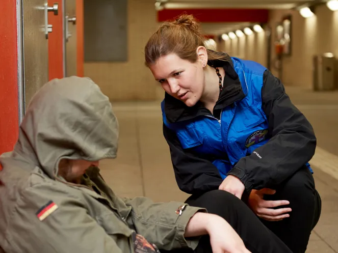 Mitarbeiterin Bahnhofsmission beugt sich herunter zu einer Person mit Kapuze ins Gesicht gezogen