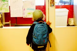 Ein kleiner Junge mit Rucksack schaut von außen durch eine geschlossene Glastür.