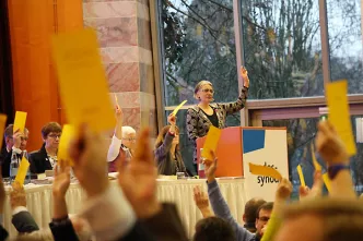 Alle Synodale auf dem Podium halten gelbe Abstimmungszettel hoch