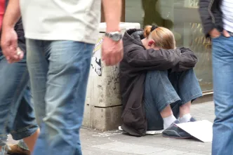 Eine junge Frau sitzt in gebückter Haltung am Boden einer belebten Einkaufsstraße, den Kopf auf den Armen vergraben. Vor ihr liegt ein Papier, möglicherweise für Spenden. Passant*innen gehen achtlos an ihr vorbei.