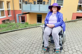 Gisela Breuhaus sitzt in lila Kleidung im Rollstuhl vor ihrem Wohnhaus in Bonn