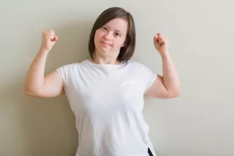 Eine Frau mit Down Syndrom hält beide Arme hoch und zeigt selbstbewusst ihre Muskeln