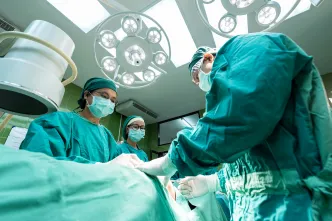 Ein Team von Chirurg*innen im Operationssaal bei einem Eingriff. Alle tragen grüne OP-Kleidung, Mundschutz und Hauben, umgeben von OP-Lampen und medizinischer Ausrüstung.