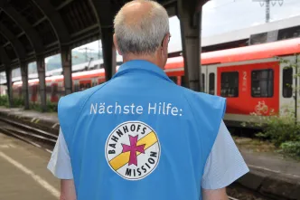 Ein Ehrenamtlicher der Bahnhofsmission steht am Bahngleis, während ein Zug vorbeifährt.
