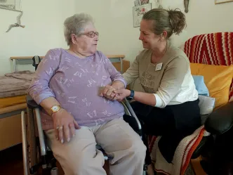 Pflegefachkraft hält die Hand einer Seniorin im Rollstuhl